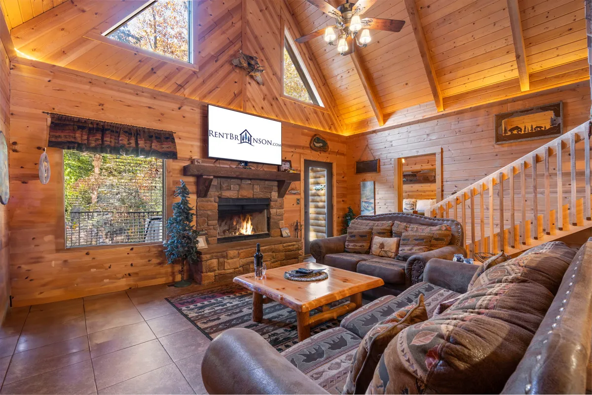 This image features a beautifully appointed living room with a striking stone fireplace as its centerpiece. The space is furnished with plush sofas and armchairs, a wooden ceiling for added warmth, and a television mounted above the fireplace. The atmosphere is cozy and inviting, perfect for gatherings, watching movies, or relaxing by the fire.