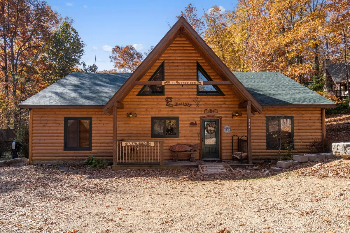 The log cabin exterior is highlighted with a sloped roof, a sturdy wooden bench, and vibrant autumn foliage, set beside an open area that hints at generous parking or maneuvering space for boats and vehicles.
