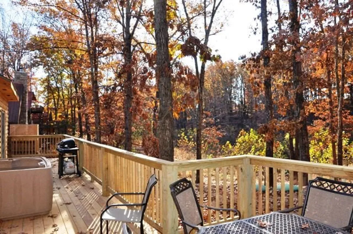 A spacious wooden deck attached to the cabin, featuring a propane grill and a bubbling hot tub nestled among tall trees. The deck is elevated with sturdy railings, blending into the surrounding natural landscape, and provides a peaceful retreat for outdoor dining and relaxation after a day on the lake.