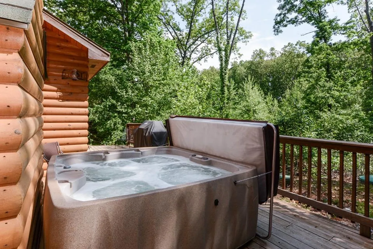 A wooden deck features a bubbling hot tub surrounded by greenery, offering a relaxing outdoor amenity just steps from the cabin. The scene suggests unwinding in nature after a lively event or concert.