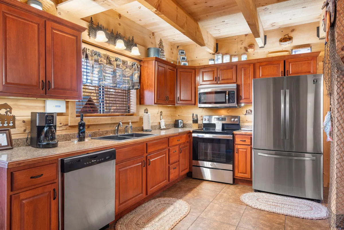 This image displays a modern, open kitchen with wooden cabinetry, stainless steel appliances, and tasteful countertop decor. Sunlight filters in through a window, illuminating the space and underscoring the kitchen’s welcoming vibe.