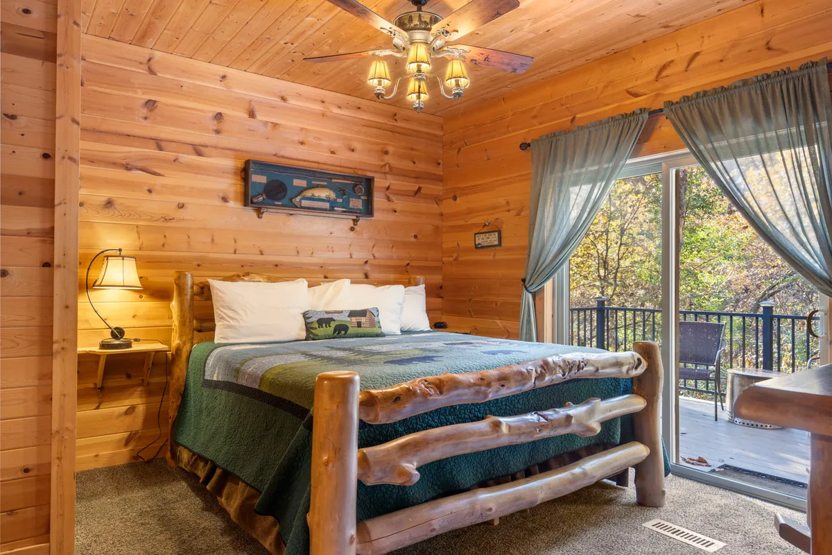 A serene king bedroom with a wooden bed frame and plaid blanket, bathed in natural light from a large window that offers a tranquil view. The decor is warm and comfortable, with an emphasis on premium linens and a restful atmosphere.
