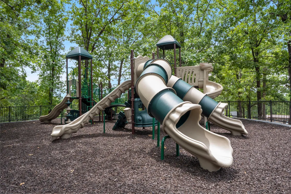 This image showcases a vibrant playground area surrounded by tall trees, with slides and climbing structures clearly visible. Families and children can be seen enjoying themselves, capturing the spirit of outdoor play and togetherness.
