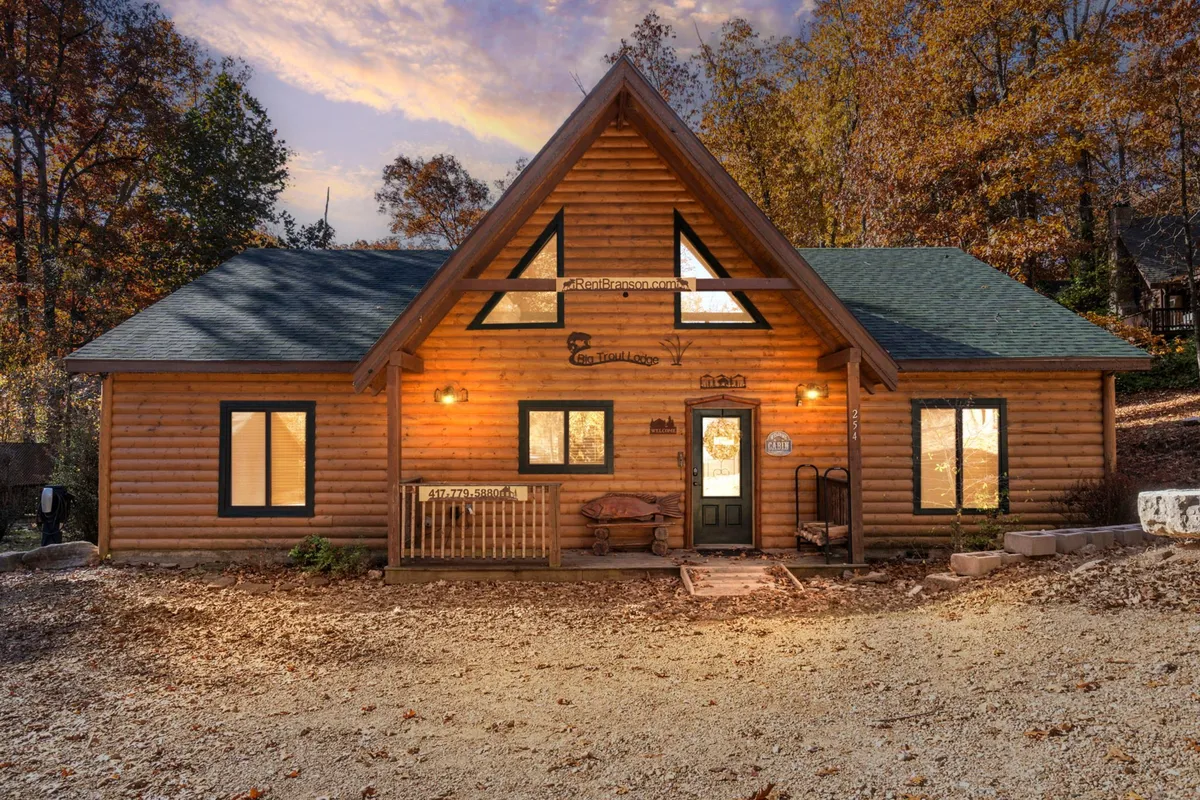 A charming log cabin with a front porch and ample seating, framed by tall trees and natural surroundings. The inviting porch offers a spot to unwind after outdoor adventures, while large windows provide glimpses of the peaceful landscape beyond.