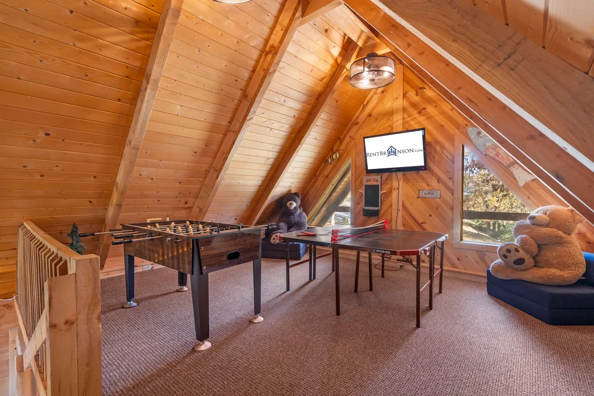 This interior image features a vibrant game loft with a foosball table, a ping pong table, a large stuffed bear, and a visible TV screen. The space is arranged for both play and relaxation, with open floor space for games and cozy touches for movie nights or lounging.