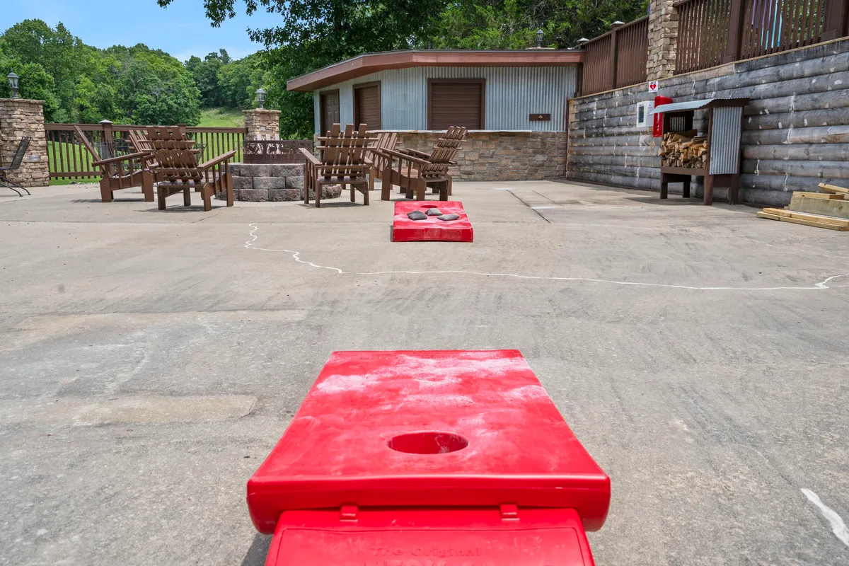 An exterior area featuring red cornhole boards set up on a patio, with wooden chairs gathered around a cozy fire pit and a stone wall structure in the background. The space is set up for outdoor games and evening gatherings, inviting guests to enjoy friendly competition and relaxed conversations by the fire.