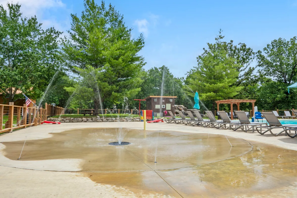 This exterior scene features a lively splash pad with water features surrounded by lounge chairs and trees. Children are seen playing joyfully in the water, while adults relax and socialize in the shade or sunbathe in the inviting atmosphere.