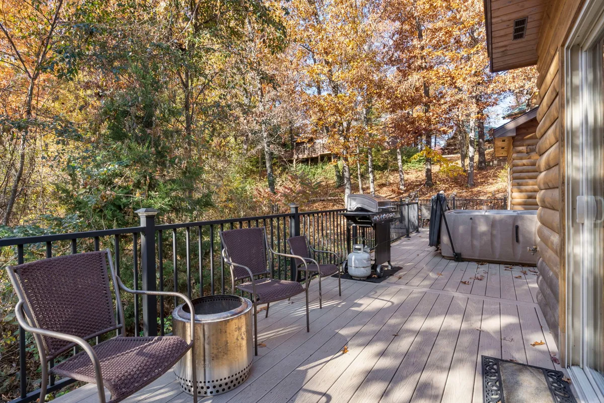 A spacious outdoor deck featuring comfortable deck chairs, a fire pit, a BBQ grill, and a bubbling hot tub, all surrounded by lush foliage. The space is designed for relaxation, outdoor dining, and social gatherings, with multiple seating options and amenities visible.